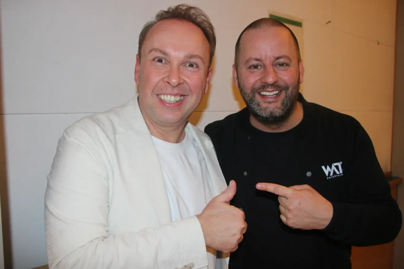 Zwei Männer lächeln und posieren für ein Foto, einer in einer weißen Jacke und der andere in einem schwarzen Shirt mit einem weißen Logo darauf.