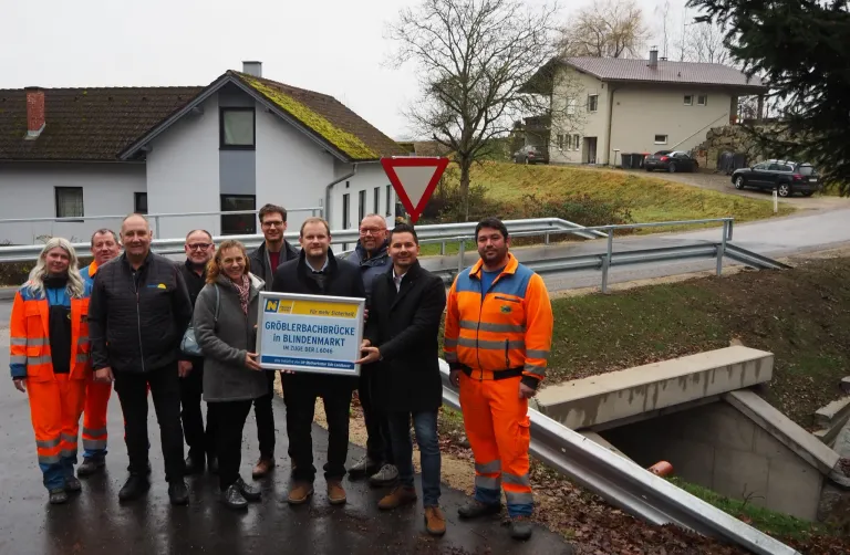 Eine Gruppe von Menschen steht auf einer Brücke mit einem Mann in orangefarbener Arbeitskleidung, der ein Schild mit der Aufschrift Groblerbachbrucke in Blindenmarkt hält.