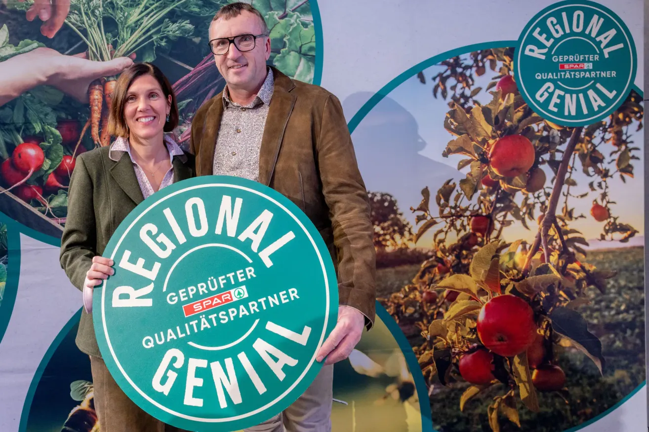Ein Mann und eine Frau lächeln und stehen hinter einem grünen Schild mit der Aufschrift 'Regional Geprüfter Qualitätspartner', mit einem Apfelbaum im Hintergrund.