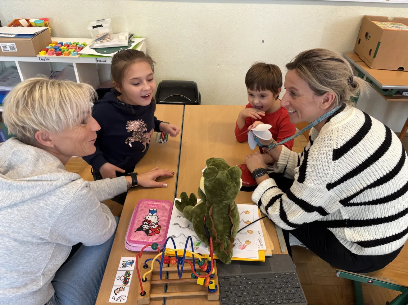Vier Personen sitzen um einen Tisch in einem Klassenzimmer. Eine Frau in Grau spricht mit einem Mädchen in Schwarz. Ein Junge in Rot hält ein Papier und eine Frau in Streifen lächelt. Ein Laptop, Spielzeug und ein Stiftetui liegen auf dem Tisch.