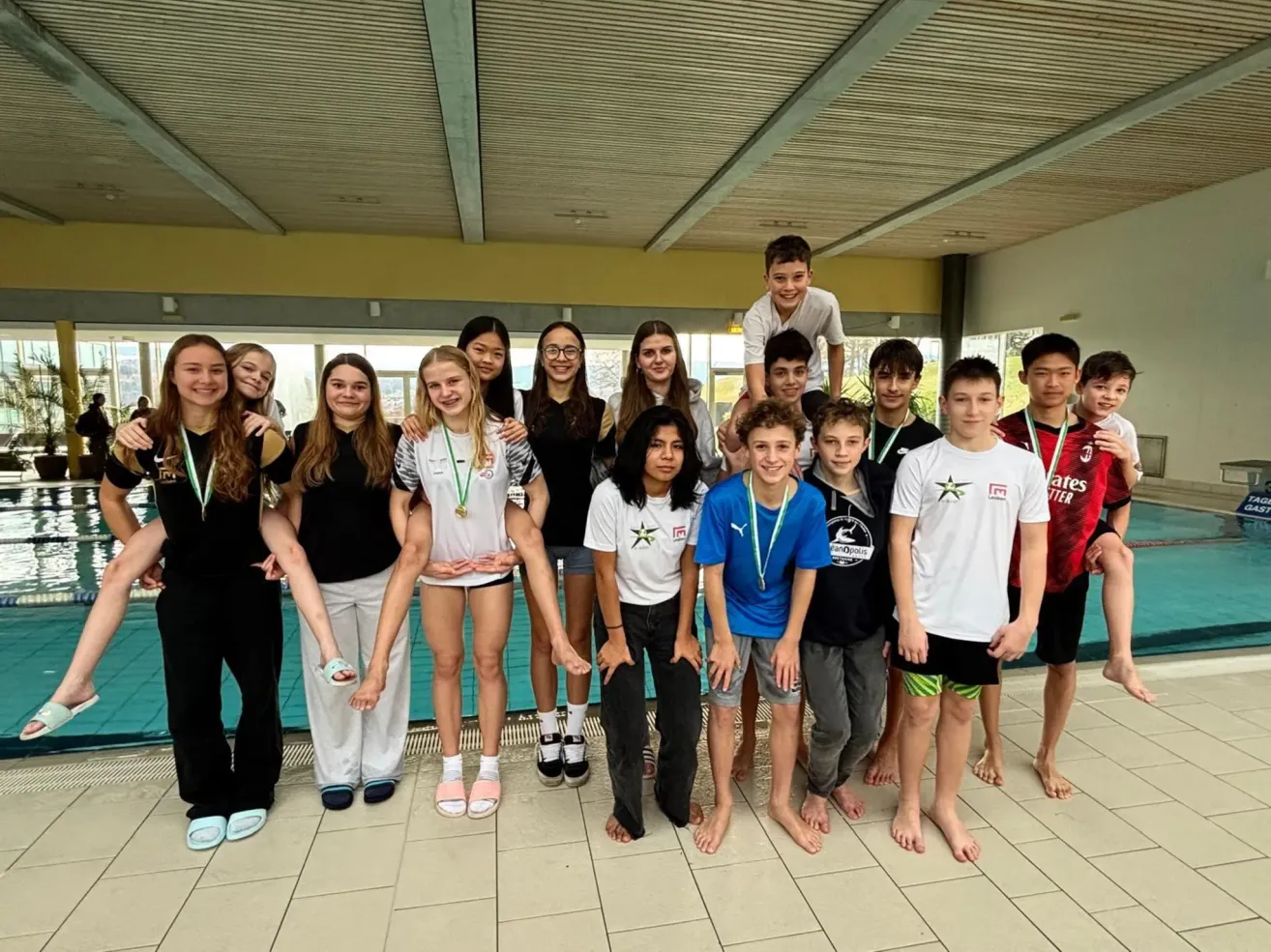 Eine Gruppe junger Schwimmer steht in einem Schwimmbereich mit Medaillen, lächelt und posiert für ein Foto. Dahinter ist ein Schwimmbecken zu sehen.