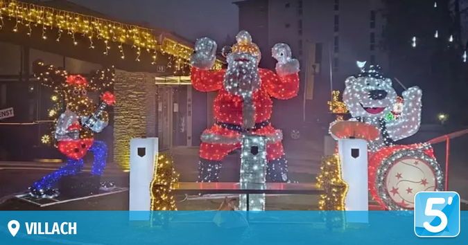 Eine Weihnachtsdekoration in einer Stadt zeigt den winkenden Weihnachtsmann, einen Schlagzeug spielenden Bären und einen Gitarristen in festlicher Kleidung, alle beleuchtet am Nachthimmel.