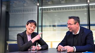 Zwei Personen sitzen an einem Tisch mit Gläsern Wasser, lächelnd. Die Frau zeigt auf etwas. Der Mann trägt eine Brille und eine Uhr. Hinter ihnen ist eine Glaswand.