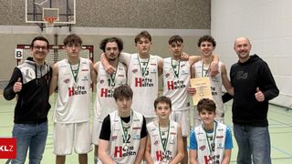 Eine Basketballmannschaft posiert für ein Foto in einer Turnhalle, alle tragen weiße Trikots mit 'Hilfe' und Medaillen um den Hals. Ein Mann hält ein Zertifikat.