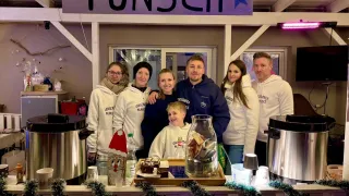 Eine Familie steht lächelnd vor einem festlichen Tresen. Hinter ihnen befindet sich ein Schild mit der Aufschrift 'Punsch'. Sie tragen passende weiße Sweatshirts. Auf dem Tresen stehen zwei große silberne Töpfe, ein großes Glasgefäß gefüllt mit Münzen und ein Tablett mit verschiedenen Leckereien. Ein Hintergrund aus Weihnachtsdekorationen verleiht dem Bild den festlichen Charakter.