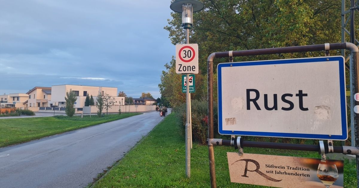 A street with a 30 km/h speed limit sign. A sign reading 'Rus' and another reading 'Südwein' is attached to a pole. Trees and a building are in the background.