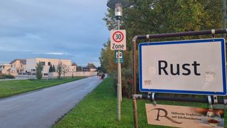 Eine Straße mit einem 30 km/h Geschwindigkeitsbegrenzungsschild. Ein Schild mit der Aufschrift 'Rus' und ein weiteres mit der Aufschrift 'Südwein' ist an einem Pfosten befestigt. Bäume und ein Gebäude sind im Hintergrund.
