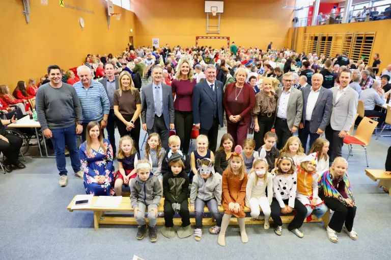 Eine große Gruppe von Erwachsenen und Kindern in Festkostümen posiert für ein Foto in einer Turnhalle, wobei einige Masken tragen und einige hinter einer Holzbank stehen.