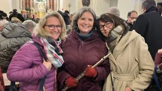 Drei Frauen stehen eng beieinander in einer Kirche und lächeln. Die Frau in der Mitte hält eine Flöte. Die Kirche ist geschmückt, und ein Bilderrahmen ist an der Wand montiert.