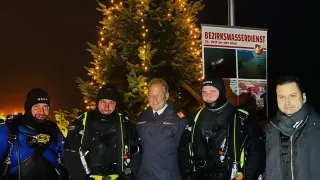 Fünf Personen stehen vor einem geschmückten Weihnachtsbaum bei Nacht. Vier tragen Taucheranzüge, einer einen Anzug. Ein Schild zeigt 'Bezirkswasserdienst'.