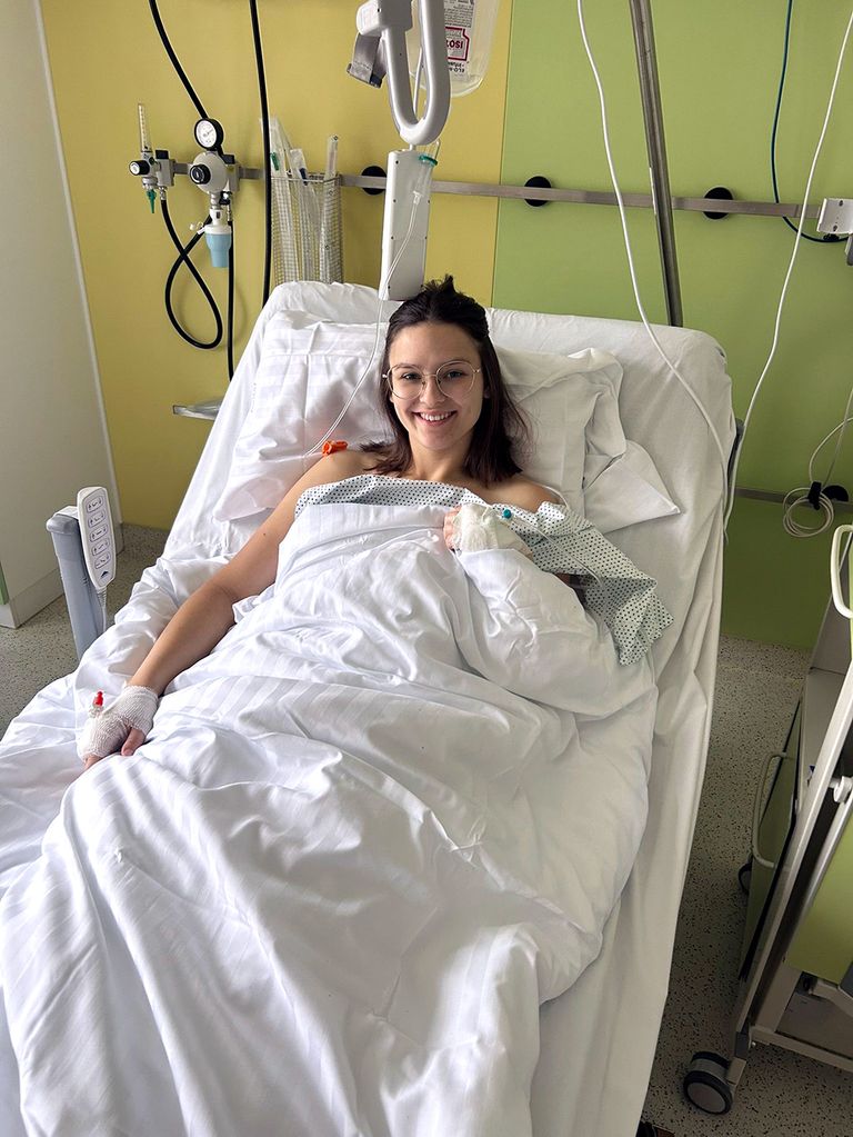 Eine Frau mit einem bandagierten Hand- und Arm liegt im Krankenhausbett und lächelt. Hinter ihr ist medizinische Ausrüstung an der Wand befestigt.