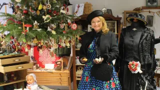 Eine Frau in einem vintage-Stil-Kleid posiert vor einem geschmückten Weihnachtsbaum und einem Regal mit verschiedenen Ornamenten und Gegenständen.