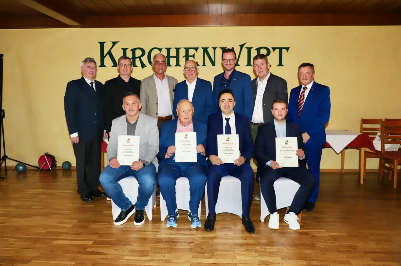 Eine Gruppe von Männern, formell gekleidet, posiert für ein Foto vor einem gelben Hintergrund, auf dem 'KIRCHEN VIRT' steht. Sie halten Urkunden in den Händen und sitzen auf weißen Stühlen.
