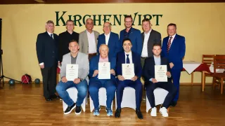 A group of men, formally dressed, are posing for a photo in front of a yellow backdrop that reads 'KIRCHEN VIRT.' They are holding certificates and seated on white chairs.