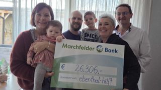 Eine Familie mit sechs Personen, darunter Kinder, hält einen großen Scheck über 26.306 Euro von der Marchfelder Bank. Sie stehen lächelnd vor einem Fenster.
