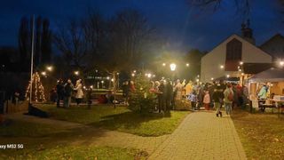 Eine Menschenmenge versammelt sich nachts in einem Park, geschmückt mit Weihnachtslichtern und Bäumen, einige stehend, andere auf Bänken sitzend.