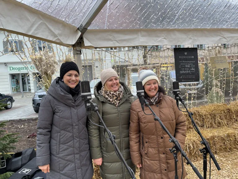 Drei Frauen stehen unter einem Baldachin, jede hält ein Mikrofon in der Hand. Sie sind warm in Wintermänteln und Mützen gekleidet. Hinter ihnen steht ein Schild mit Text in einer Fremdsprache und im Hintergrund ein Gebäude mit leuchtenden Lichtern.