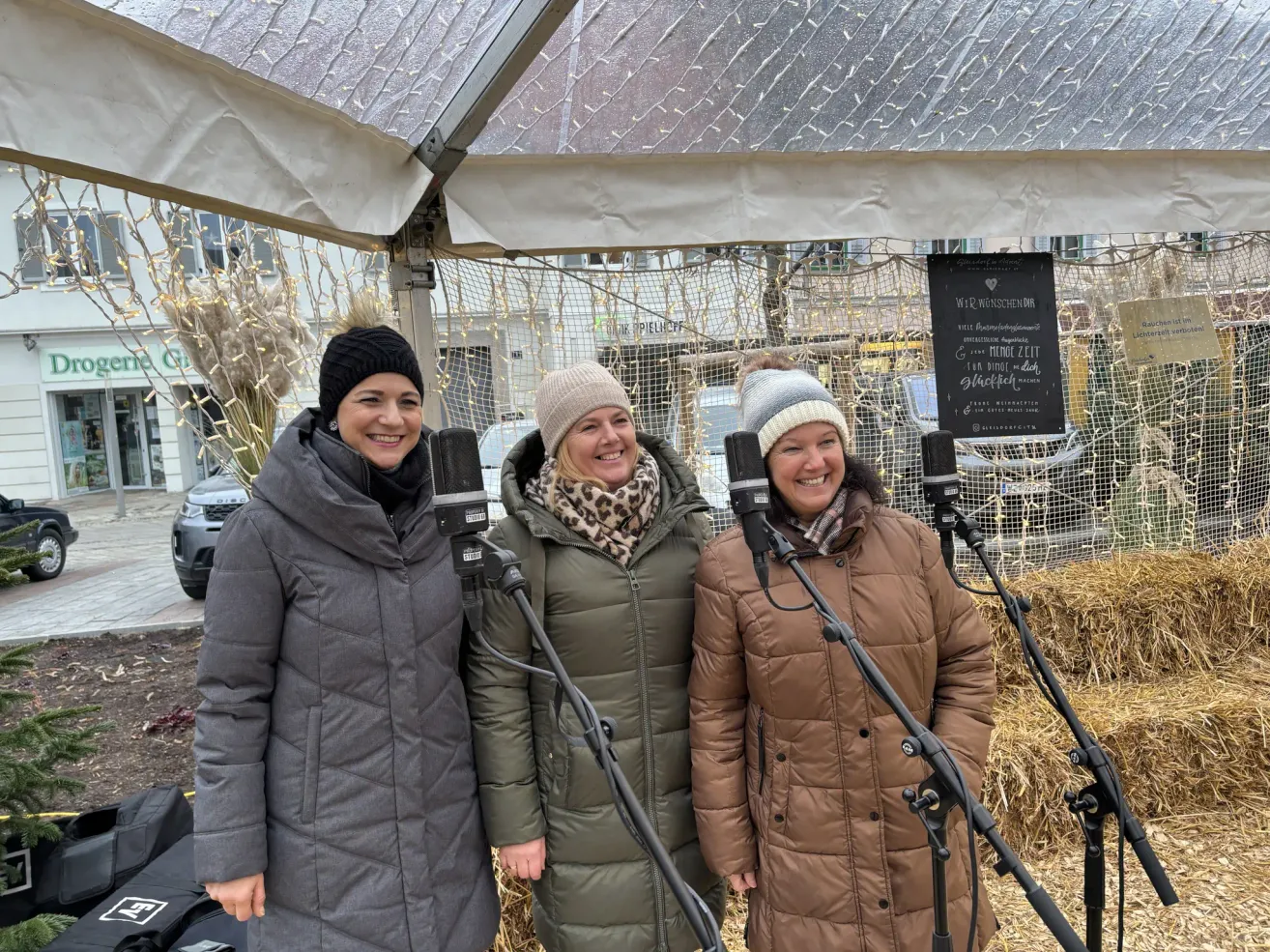 Drei Frauen stehen unter einem Baldachin, jede hält ein Mikrofon in der Hand. Sie sind warm in Wintermänteln und Mützen gekleidet. Hinter ihnen steht ein Schild mit Text in einer Fremdsprache und im Hintergrund ein Gebäude mit leuchtenden Lichtern.