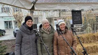 Drei Frauen stehen unter einem Baldachin, jede hält ein Mikrofon in der Hand. Sie sind warm in Wintermänteln und Mützen gekleidet. Hinter ihnen steht ein Schild mit Text in einer Fremdsprache und im Hintergrund ein Gebäude mit leuchtenden Lichtern.