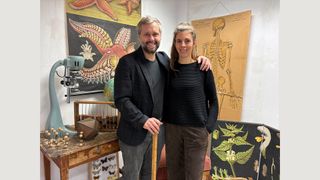 Ein Mann und eine Frau stehen gemeinsam in einem wissenschaftlichen Raum, lächeln für die Kamera. Der Mann hält einen Stock und beide sind von verschiedenen wissenschaftlichen Illustrationen und Objekten umgeben.