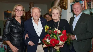 Vier Menschen stehen zusammen in einem Raum. Ein Mann und eine Frau halten einen Blumenstrauß, während eine Frau mit Brille links steht.