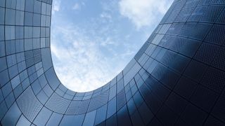 Ein modernes Gebäude mit geschwungener Form, das den blauen Himmel mit einigen Wolken reflektiert. Die Struktur besteht aus Metallplatten mit einem gitterartigen Muster.