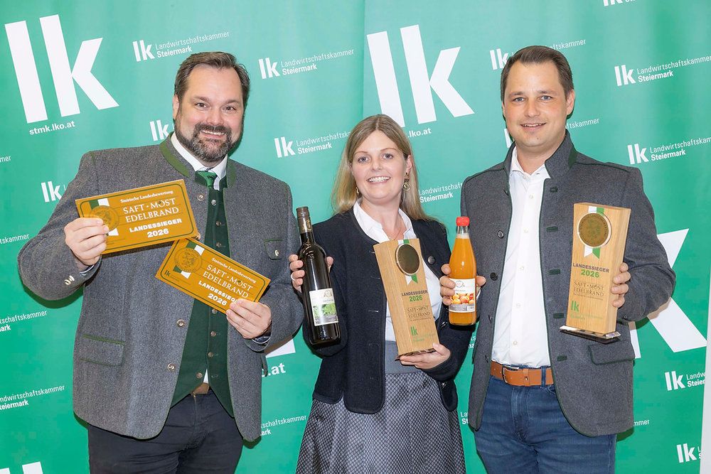 Drei Personen stehen gemeinsam vor einem grünen Hintergrund. Der Mann auf der linken Seite hält eine Flasche Wein, die Frau in der Mitte hält eine Flasche Saft und der Mann auf der rechten Seite hält eine Plakette. Sie alle lächeln und posieren für ein Foto.