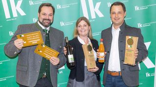 Drei Personen stehen gemeinsam vor einem grünen Hintergrund. Der Mann auf der linken Seite hält eine Flasche Wein, die Frau in der Mitte hält eine Flasche Saft und der Mann auf der rechten Seite hält eine Plakette. Sie alle lächeln und posieren für ein Foto.