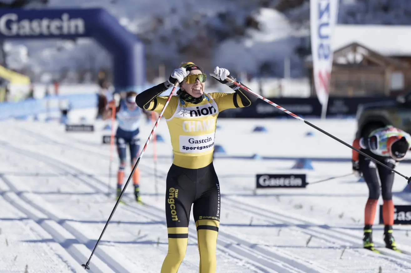 Eine Skifahrerin in einem gelben und schwarzen Outfit steht im Schnee mit Skistöcken. Hinter ihr ist ein weiterer Skifahrer, und es gibt Banner und Schilder im Hintergrund.