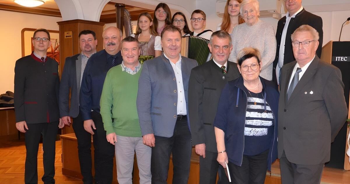 Eine Gruppe von Menschen steht lächelnd für ein Foto zusammen, mit einer hölzernen Wand und einer Harfe im Hintergrund.