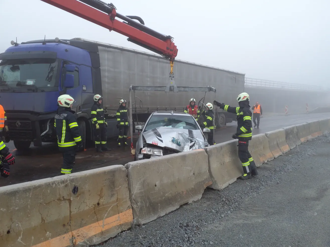 Einsatzkräfte arbeiten daran, ein beschädigtes Auto von einer Autobahn zu entfernen. Ein Kran ist am Auto befestigt, während mehrere Feuerwehrleute in der Nähe stehen.