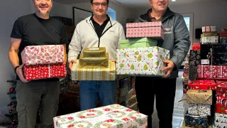 Drei Männer stehen lächelnd und halten verpackte Weihnachtsgeschenke. Ein Mann hält ein Geschenk mit rotem und weißem Muster, ein anderer ein Geschenk mit goldenem Muster und der dritte ein Geschenk mit grünem Muster. Sie stehen vor einem Haufen Weihnachtsgeschenke.