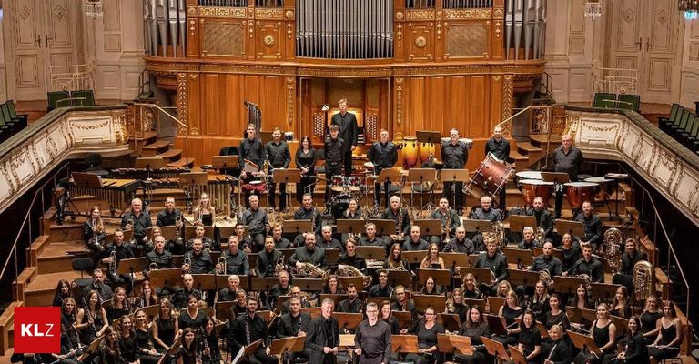 Ein großes Orchester befindet sich in einem Konzertsaal, mit Musikern in schwarzer Kleidung und Orchesterständen vor ihnen. Einige stehen, während andere sitzen, und alle schauen in Richtung des Dirigenten. Hinter dem Orchester befindet sich eine Großorgel und eine Treppe.