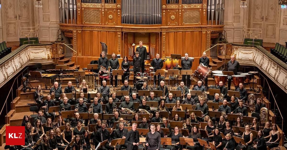 Ein großes Orchester befindet sich in einem Konzertsaal, mit Musikern in schwarzer Kleidung und Orchesterständen vor ihnen. Einige stehen, während andere sitzen, und alle schauen in Richtung des Dirigenten. Hinter dem Orchester befindet sich eine Großorgel und eine Treppe.