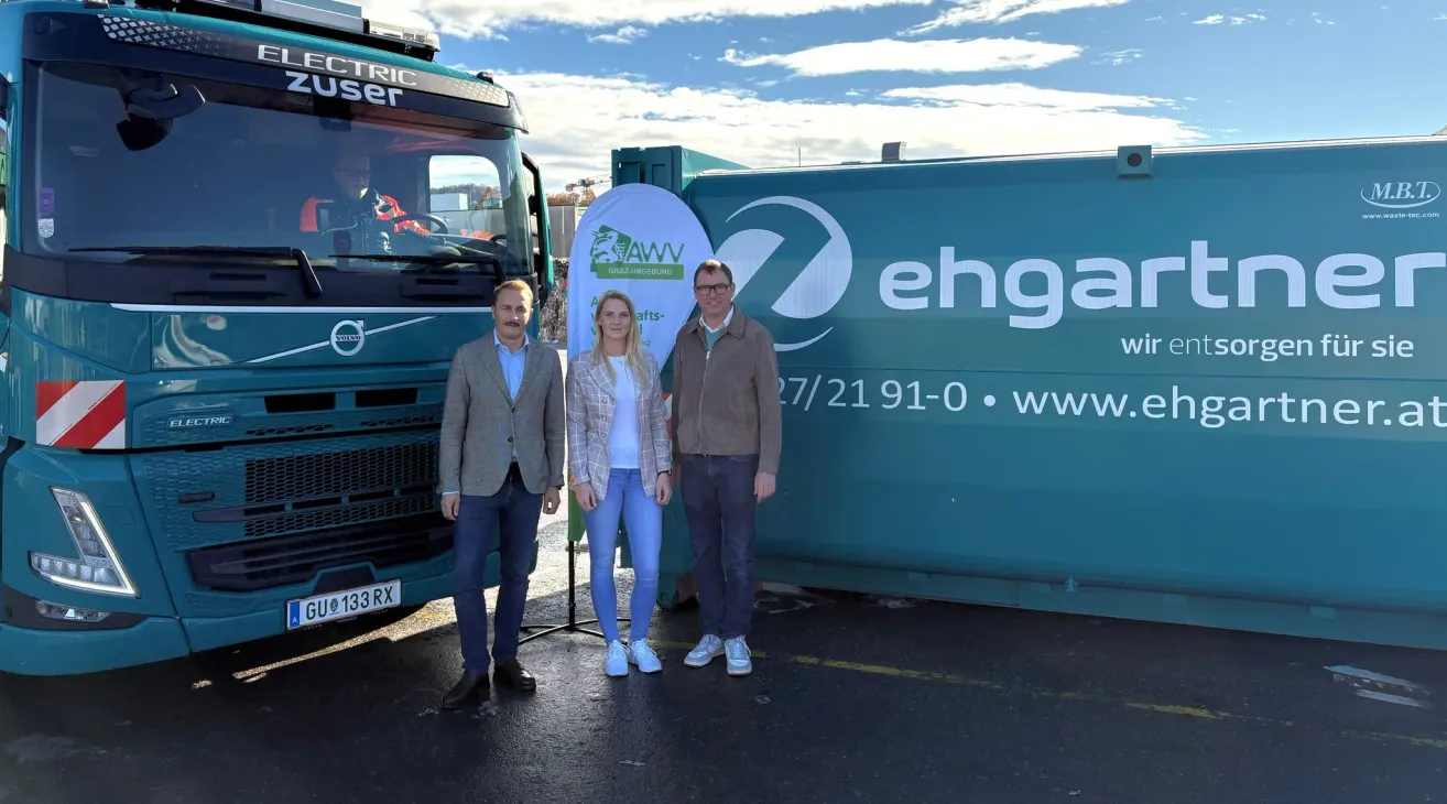 Drei Personen stehen vor einem Elektrofahrzeug, zwei Männer und eine Frau, alle lächeln. Dahinter steht ein großer grüner Container mit dem Text 'ehga'.