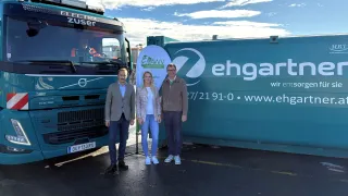 Drei Personen stehen vor einem Elektrofahrzeug, zwei Männer und eine Frau, alle lächeln. Dahinter steht ein großer grüner Container mit dem Text 'ehga'.
