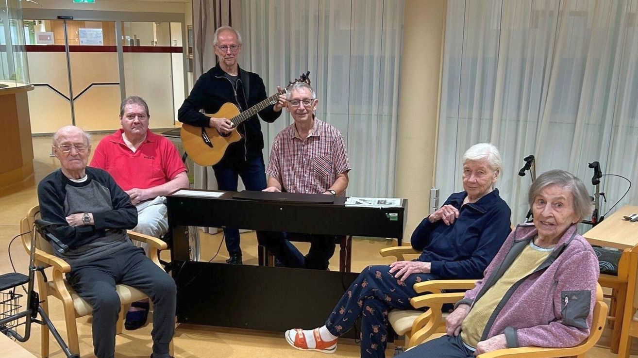 Fünf ältere Männer sitzen um ein Klavier herum. Einer spielt Gitarre dahinter. Ein anderer sitzt mit hochgelegten Füßen am Klavier. Zwei weitere sitzen an seinen Seiten.