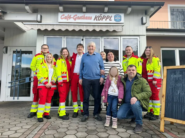 Eine Gruppe von Menschen in Sicherheitsanzügen und einem Mann in einem blauen Pullover posiert für ein Foto vor einem Gebäude mit einem Schild, das 'Cafe-Gasthhaus Kopl' steht.