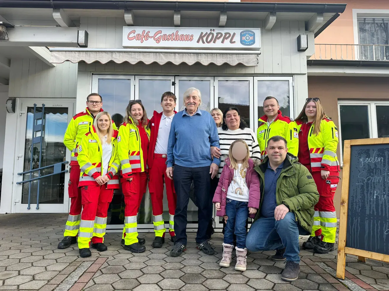 Eine Gruppe von Menschen in Sicherheitsanzügen und einem Mann in einem blauen Pullover posiert für ein Foto vor einem Gebäude mit einem Schild, das 'Cafe-Gasthhaus Kopl' steht.