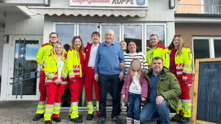 Eine Gruppe von Menschen in Sicherheitsanzügen und einem Mann in einem blauen Pullover posiert für ein Foto vor einem Gebäude mit einem Schild, das 'Cafe-Gasthhaus Kopl' steht.