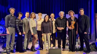 Eine Gruppe von Musikern steht auf einer Bühne mit einem blauen Vorhang im Hintergrund. Eine Frau in der Mitte hält eine Geige.