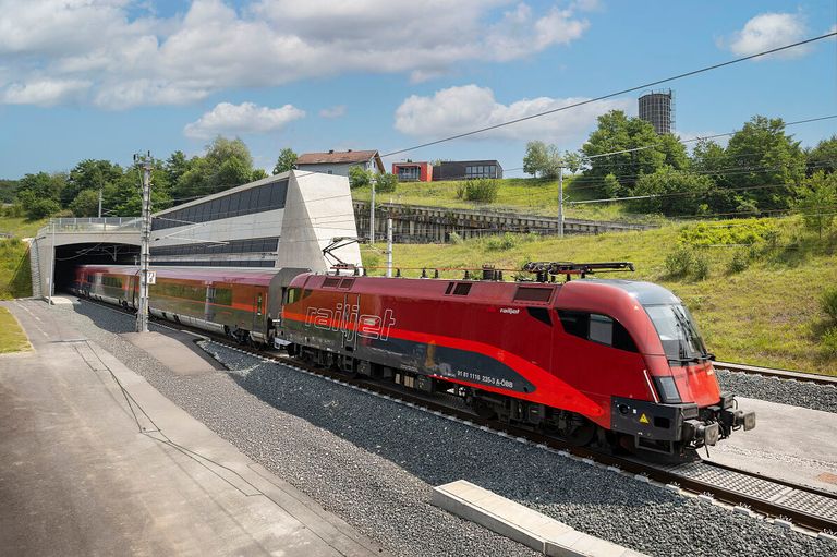 Ein roter Zug mit 'railjet' auf der Seite steht auf den Gleisen und ist an einem Bahnhof mit einer Betonplattform geparkt.