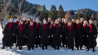 Ein Chor von Erwachsenen in schwarzen Mänteln mit roten Schals steht im Schnee mit Bäumen und Bergen im Hintergrund.
