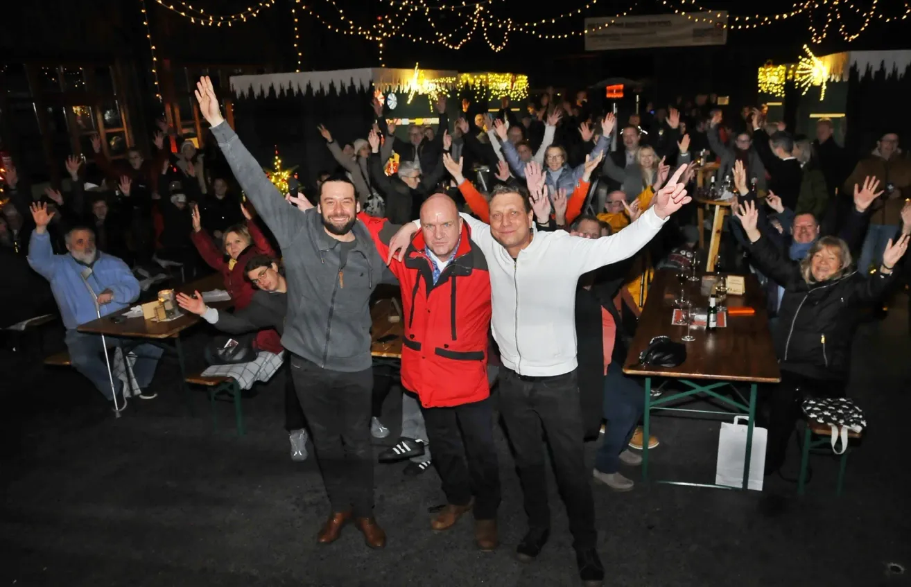 Drei Männer feiern zusammen und heben ihre Arme bei einer festlichen Veranstaltung im Freien mit einer Menschenmenge, Lichterketten und Tischen mit Gläsern.