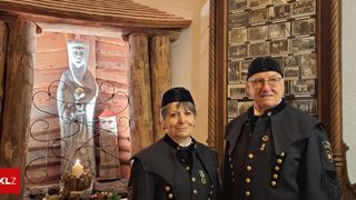 Zwei Personen in traditionellen Uniformen stehen nebeneinander. Dahinter befindet sich eine Holzstruktur mit einer Statue und einer Kerze.