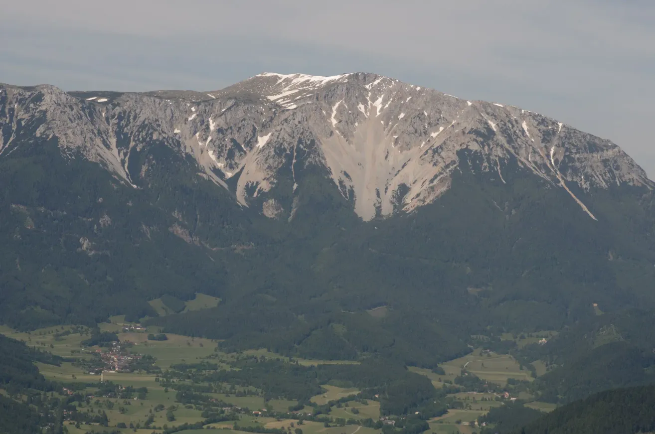 Luftaufnahme eines schneebedeckten Berges mit einem weitläufigen grünen Tal darunter, unter einem teilweise bewölkten Himmel.