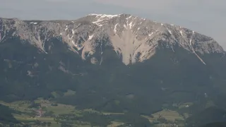 Aerial view of a snow-capped mountain with a vast green valley below, under a partly cloudy sky.