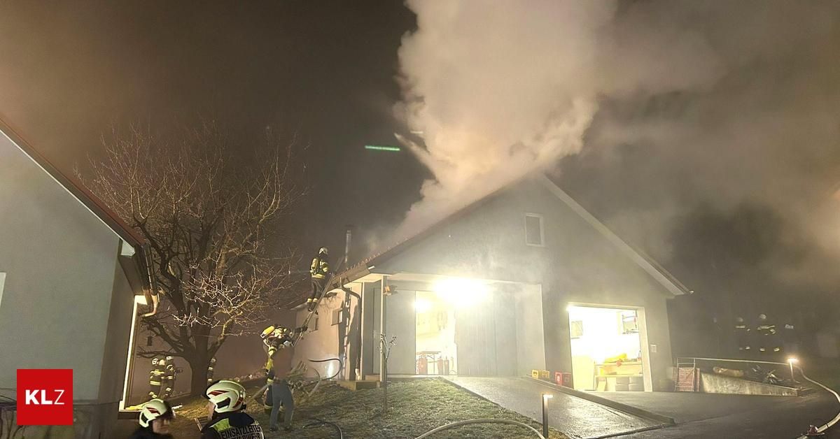 Feuerwehrleute stehen bei Nacht auf dem Dach eines Hauses und bekämpfen einen Brand, bei dem Rauch aus dem Schornstein aufsteigt. Sie sind mit Helmen und Schutzausrüstung ausgestattet.