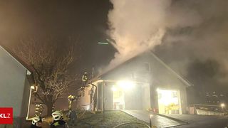 Feuerwehrleute stehen bei Nacht auf dem Dach eines Hauses und bekämpfen einen Brand, bei dem Rauch aus dem Schornstein aufsteigt. Sie sind mit Helmen und Schutzausrüstung ausgestattet.