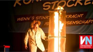 Eine Person in einem weißen Laborkittel steht auf der Bühne und hält eine große, zylindrische Fackel mit einem brennenden Feuer im Inneren. Der Hintergrund zeigt ein Banner mit den Worten Wenn's Stink Is und einer leuchtenden Flamme.